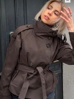Mörkbrun trenchcoat med knytskärp - Trenchcoat från  Bershka i storlek L som jag hann köpa men nu hittat en annan vårjacka efter jag hann dra av lappen:/ kollar därför om det finns intresse här. Stilren mörkbrun trenchcoat med dubbelknäppning, höga axelklaffar och knytskärp i midjan. Jackan har stora knappar och hög krage som kan stängas upp. Tillverkad i ett kraftigt tyg som ger en snygg siluett.
