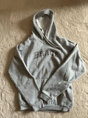 Grå hoodie med huva - Säljer en grå hoodie med huva och magficka. Lite osäker på märket vet inte ifall det är Brace som det står på. Skön med snyggt tryck.💕