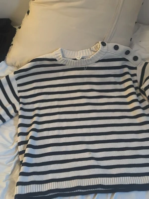 Randig stickad tröja med knappar - Säljer en stickad tröja med svarta och vita ränder. Tröjan har rund halsringning och fyra stora knappar på ena axeln som snygg detalj. Perfekt tröja inför sommaren och passar till allt!!