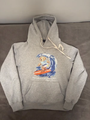 Grå  Polo Björn Ralph Lauren hoodie - Säljer en grå hoodie från Ralph Lauren med den ikoniska Polo Bear som surfar på en våg framtill. Hoodien har huva med dragsko, magficka och ribbade muddar. Trycket är färgglatt med blått, orange och vitt. Perfekt för dig som gillar streetwear och snygga detaljer. PRIS KAN DISKUTERAS. STÅR ATT DEN ÄT M MEN PASSR MELLAN M-S