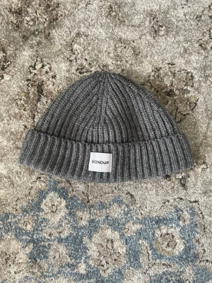 Dondup mössa - Säljer en stilren grå ribbstickad mössa från Dondup. Mössan har uppvikt kant och en vit patch med Dondup-loggan framtill. Tillverkad i mjukt och varmt material som håller dig bekväm under kalla dagar. Enkel och clean design som passar till det mesta.