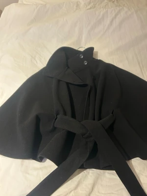 Svart cape-kappa från Gina Tricot - Stilren svart cape-kappa från Gina Tricot med hög krage, knappar och knytskärp i midjan. Kappan har en elegant, vid siluett och är tillverkad i ett mjukt tyg som ger en snygg look. Perfekt för dig som vill ha något trendigt och bekvämt.