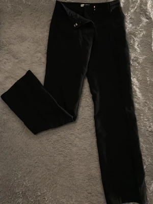 Svarta vida kostymbyxor - Säljer ett par svarta kostymbyxor med medel midja och raka, vida ben. Byxorna har klassisk design med knapp och dragkedja framtill. Perfekta för en clean och stilren look.
