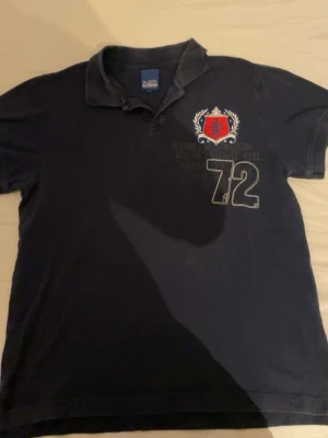 Marinblå piké med tryck och emblem - Säljer en marinblå pikétröja med krage och korta ärmar. På bröstet finns ett rött och vitt emblem samt siffran 72 tryckt i vitt. Tröjan är i bomull och har klassisk pikélook, perfekt för en sportig och avslappnad stil.