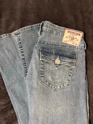 Ljusblå True Religion jeans - Säljer ett par ljusblå jeans från True Religion med klassisk kontrastsöm och ikonisk fickdetalj bak. Jeansen har normal midja, raka ben och är gjorda i mjuk denim med snygg tvätt. Perfekta för en avslappnad och trendig look. Storlek 26, joey. Knappt använda. Säljer för dom är en storlek för stor