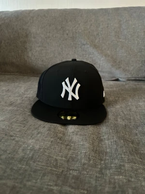 Svart New Era Yankees keps - Säljer en svart New Era keps med New York Yankees-logga broderad i vitt framtill. Klassisk flat brim och MLB-logga baktill. Kepsen är gjord i bomull och polyester, perfekt för dig som vill ha en stilren och sportig look.