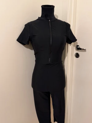 Svart jumpsuit med dragkedja - Snygg svart jumpsuit med korta ärmar och hög krage. Dragkedja framtill som går att öppna upp för en mer avslappnad look. Tight passform och stretchigt material som ger en sportig vibe. Perfekt för dig som gillar stilrena och minimalistiska plagg.