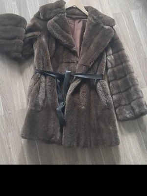 Brun fuskpäls kappa med bälte - Snygg brun fuskpäls kappa med bred krage och svart knytbälte i midjan. Kappan har en klassisk, rak passform och är helfodrad för extra komfort. Perfekt för dig som vill ha en trendig och varm look under kalla dagar.