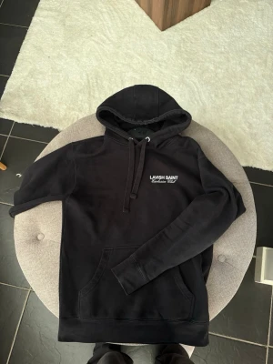 Svart hoodie från Lavish Saint - Svart hoodie från Lavish Saint med vit text på bröstet och ryggen. Klassisk modell med huva, dragsnören och känguruficka framtill. Mjuk bomullskänsla och stilren design, perfekt för en avslappnad streetwear-look.