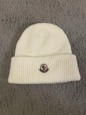 Vit ribbstickad mössa ej Moncler - Säljer en stilren vit ribbstickad mössa inte Moncler med uppvikt kant och klassisk Moncler-logga framtill. Mössan är tillverkad i mjukt material och har en clean look som passar perfekt till kalla dagar. Ej riktig
