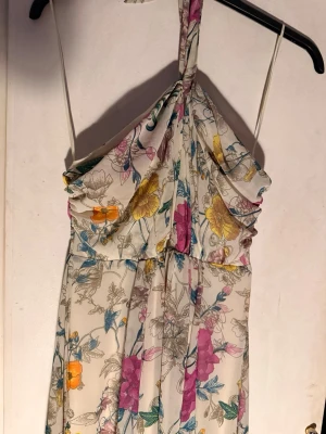 Blommig långklänning med halterneck - Säljer en romantisk långklänning med halterneck och draperad överdel. Klänningen har ett färgglatt blommönster i rosa, gul, orange och blå på en ljus botten. Perfekt för sommarens alla tillfällen. Materialet känns lätt och luftigt.