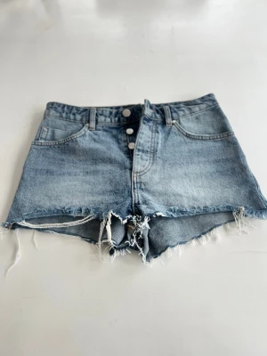 Blå jeansshorts med fransig kant - Säljer ett par klassiska blå jeansshorts med hög midja och rå, fransig kant. Shortsen har knappar framtill, femficksmodell och en avslappnad passform. Perfekta för sommaren och lätta att matcha med allt.