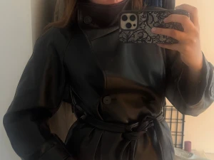 Svart trenchcoat i fuskläder - Snygg svart trenchcoat i fuskläder med dubbelknäppning och bälte i midjan. Jackan har hög krage med knappdetalj och raka ärmar. Perfekt för dig som vill ha en trendig och stilren look. OBS! En knapp har varit sönder men den är tillbaka sydd så det är inget som syns så tydligt. Den är i storlek XS men passar även S då den är oversized. Jag har vanligtvis S i storlek och den sitter oversized på mig. Kan sänkas i pris vid snabb affär!💕