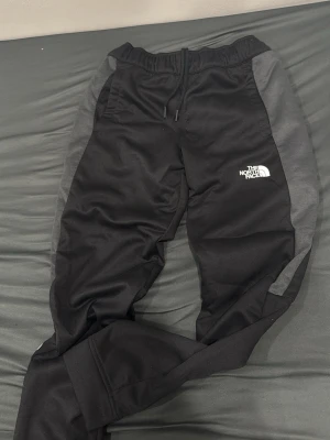Svarta mjukisbyxor från The North Face - Säljer ett par svarta mjukisbyxor från The North Face med gråa paneler på sidorna. Byxorna har resår i midjan, dragsko och logga på benet. Perfekta för chill eller träning, med en sportig och avslappnad look.