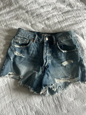 Blå slitna jeansshorts - Säljer ett par blå jeansshorts med slitna detaljer och fransig kant. Shortsen har knappgylf och råa avslut. Perfekta för sommaren. 