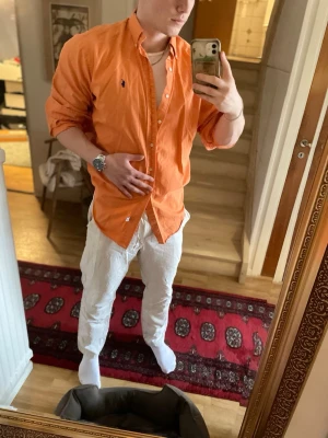 Orange skjorta från Ralph Lauren - Snygg orange skjorta från Ralph Lauren i 100% bomull. Klassisk modell med knappar framtill och liten broderad logga på bröstet. Perfekt för dig som vill sticka ut med färg och ändå hålla det stilrent.