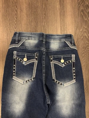 Blå bootcut jeans med stjärndetaljer - Snygga blå bootcut jeans med slitningar och dekorativa bakfickor med stjärnmönstrad kant och silvriga knappar. Jeansen har normal midja, bälteshällor och kontrastsömmar. Perfekta för dig som vill sticka ut med coola detaljer.