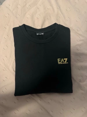 EA7 sweatshirt  - Snygg svart t-shirt från EA7 Emporio Armani med gul logga på bröstet. Klassisk rund halsringning och stilren design som passar till allt. Tillverkad i mjuk bomull för skön känsla hela dagen.