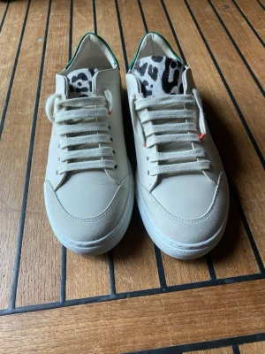 Vita sneakers Axel Arigato djurmönster - Snygga vita sneakers från Axel Arigato med svartvitt djurmönster på plösen och grön detalj på hälen. Skorna har vita snören och diskreta guldfärgade logotyper på sidan. Tillverkade i läder med klassisk låg siluett.