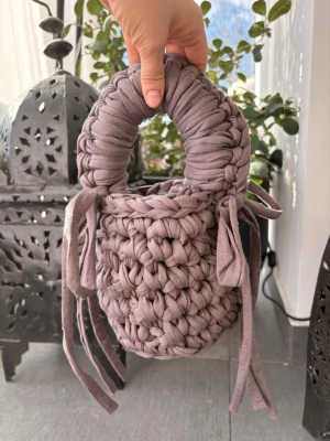 Handvirkad lila bucket bag - Unik handvirkad handväska i lila tyg med chunky struktur och cylinderform. Väskan har ett tjockt handtag och dekorativa knytband på sidorna. Perfekt för dig som gillar statement-accessoarer och vill sticka ut med en kreativ look.