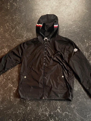 Svart vindjacka från Moncler - Snygg svart vindjacka från Moncler med huva och dragkedja framtill. Jackan har Moncler-logga på ärmen och en huva med röd och vit rand. Två fickor med tryckknappar och lätt, glansigt material som står emot vind.