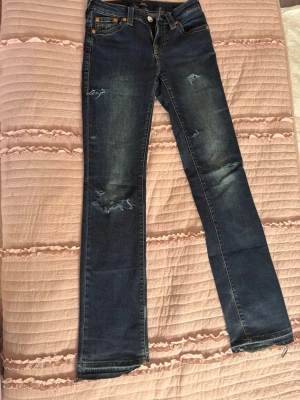 Blå slitna jeans från True Religion - Säljer ett par blå jeans från True Religion med raka ben och slitningar på både fram- och baksida. Klassisk femficksmodell med tydliga kontrastsömmar och ikonisk bakficka med broderad logga. Tillverkade i denim med snygg tvätt och detaljer.