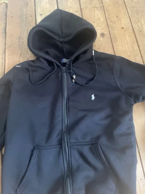 Svart set från Polo Ralph Lauren - Säljer ett svart set från Polo Ralph Lauren med hoodie och matchande byxor. Hoodien har dragkedja, fickor och justerbar huva. Byxorna har resår i midjan och dragsko. Båda plaggen har den klassiska Polo-loggan broderad på bröstet och benet. Mjukt material, perfekt för chill.