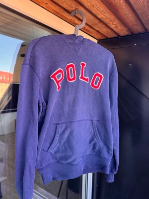 Marinblå hoodie från Polo Ralph Lauren🤩🙌 - Mörkblå hoodie från Polo Ralph Lauren med stor röd och vit POLO-text på bröstet. Klassisk känguruficka framtill och ribbade muddar vid ärmar och nederkant. Tillverkad i mjukt bomullsmaterial, perfekt för en avslappnad streetwear-look.🙌