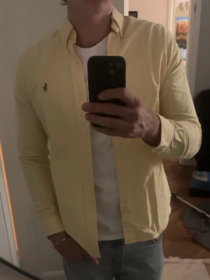 Gul skjorta från Ralph Lauren - Klassisk gul skjorta från Ralph Lauren med lång ärm och knappstängning framtill. Skjortan har den ikoniska broderade loggan på bröstet och är tillverkad i mjuk bomull. Perfekt för dig som vill ha en stilren och fräsch look. Bröstmått: 46 cm Längd: 68 cm OBS; sista bilden visar en fläck, knappt synligt, men för ett känsligt öga. 