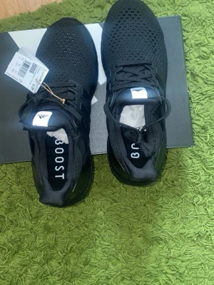 Svarta Adidas Ultraboost sneakers - Säljer ett par helsvarta Adidas Ultraboost sneakers med stickad ovandel och klassiska tre ränder på sidan. Skorna har en chunky sula i svart och snörning framtill. Perfekta för dig som gillar stilrena och bekväma sneakers med sportig vibe.
