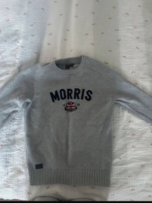 Grå stickad tröja från Morris - Snygg grå stickad tröja från Morris med mörkblå text och ett broderat brittiskt flaggmärke på bröstet. Tröjan har rund halsringning, ribbade muddar och nederkant samt en svart etikett nertill med texten 'Morris Orange'. Perfekt för en stilren look.