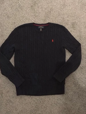 Mörkblå kabelstickad tröja Polo Ralph Lauren - Snygg mörkblå kabelstickad tröja från Polo Ralph Lauren med rund halsringning och den klassiska röda logotypen broderad på bröstet. Tröjan har långa ärmar och ribbade muddar vid ärmslut och nederkant. Perfekt för en stilren och avslappnad look. Tröjan är strl 158 men sitter xs 