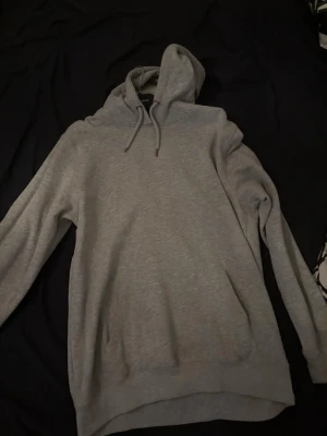 Grå basic hoodie med huva - En stilren grå hoodie från Jack & Jones med klassisk huva och dragsnören. Hoodien har en avslappnad passform, lång ärm och en stor magficka framtill. Perfekt för en chill och enkel look. Tillverkad i mjukt bomullsmaterial.