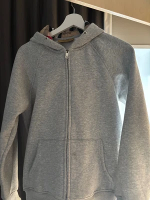 Grå zip hoodie med huva - Klassisk grå hoodie med dragkedja framtill och stor ficka på magen. Hoodien har en mjuk och skön känsla, ribbade muddar vid ärmslut och nederkant samt en rymlig huva. Perfekt för en avslappnad och stilren look.