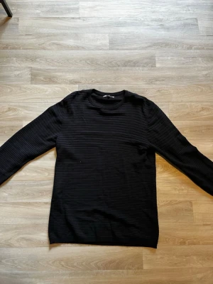 Svart ribbstickad tröja - En stilren svart stickad tröja med diskreta horisontella ribbor. Tröjan har rund halsringning och långa ärmar, perfekt för lager på lager. Passar dig som gillar minimalistisk och klassisk stil.