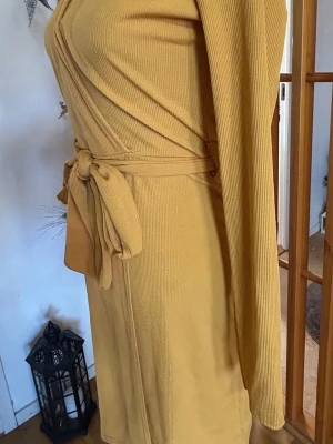 Gul omlottklänning från NLY Trend - Snygg gul omlottklänning från NLY Trend i ribbad polyester. Klänningen har lång ärm, v-ringad hals och ett knytband i midjan som ger en schysst siluett. Kort längd och mjukt, stretchigt material som sitter bekvämt. Perfekt för dig som gillar färg och stil.