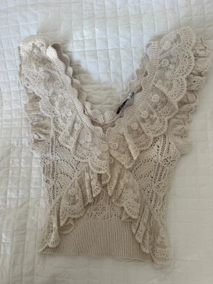 Beige spetsprydd stickad topp - Supersöt beige stickad topp med breda volangdetaljer i spets längs v-ringningen och axlarna. Toppen är ärmlös i storlek S. Priset kan diskuteras 💞