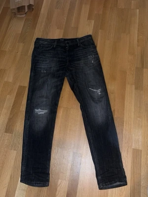Svarta slitna jeans med raka ben - Säljer ett par svarta jeans med slitningar och raka ben. Jeansen har klassisk femficksmodell och snygga slitdetaljer på framsidan för en trendig look. Perfekta för dig som gillar en avslappnad och cool stil.