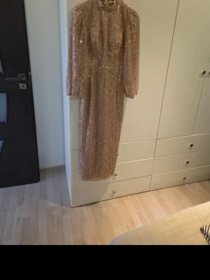 Glittrig lång klänning med paljetter - Säljer en lång, beige klänning täckt av glittrande paljetter. Klänningen har långa ärmar, hög hals och rak passform. Materialet är skimrande och ger en lyxig känsla. Perfekt för dig som vill sticka ut med en elegant look.