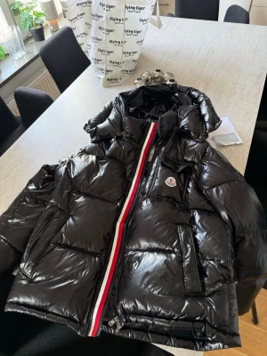 Moncler jacka - Säljer en svart glansig pufferjacka från Moncler med ikonisk logga på bröstet. Jackan har en röd, vit och blå rand längs dragkedjan, stora fickor och en rejäl huva. Perfekt för kalla vinterdagar och riktigt snygg streetstil.