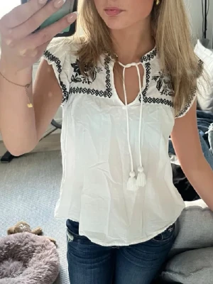 Vit blus med broderi och knyt - Söt vit blus med svart broderat mönster vid axlarna och halsringningen. Blusen har korta ärmar, knytband med tofsar framtill och en lätt, luftig passform. Perfekt för dig som gillar boho-vibbar och detaljer.