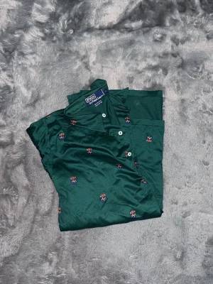 Polo Ralph Lauren All-Over Crest Embroidery - En extremt snygg mörkgrön pikétröja från Polo Ralph Lauren med ett sällsynt och detaljrikt mönster. Tröjan är helt täckt med små broderade vapensköldar (crests). Det här är inget man ser varje dag! • Märke: Polo Ralph Lauren • Färg: Mörkgrön (Skogsgrön) med broderier i rött, blått och gult. • Mönster: All-over crest embroidery (broderade vapensköldar). • Passform: Klassisk piképassform. Sitter snyggt och stabilt. 100% bomull