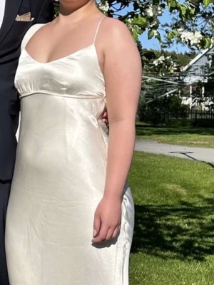 Champagnefärgad satinklänning med smala band - Säljer en elegant champagnefärgad långklänning i satin med smala axelband och öppen rygg med korsade band. Klänningen har en slits bak och en mjukt figurnära passform som ger en lyxig känsla. Perfekt för balen! Jag är en kurvig tjej och den satt perfekt, kommer vara skrynklig vid leverans då det är satin men går att stryka försiktigt! hör av dej!❤️använd endast en gång