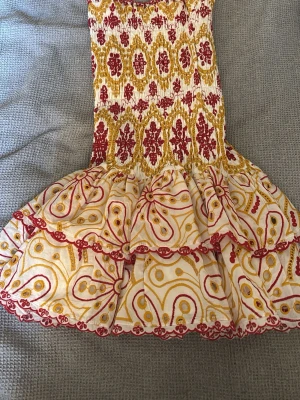 Mönstrad kortklänning från Design by Amor - Supersöt kortklänning från Design by Amor med volangkant och smock upptill. Klänningen har ett färgstarkt mönster i rött och gult på vit botten, samt fina broderade detaljer längs kanterna. Perfekt för dig som gillar unika och lekfulla plagg!