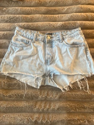 Ljusblå denimshorts med fransar - Säljer ett par ljusblå denimshorts från Lager 157 med råa, fransiga kanter och klassisk femficksdesign. Shortsen har låg midja och en avslappnad passform, perfekta för varma dagar. Stängs med knapp och dragkedja framtill.