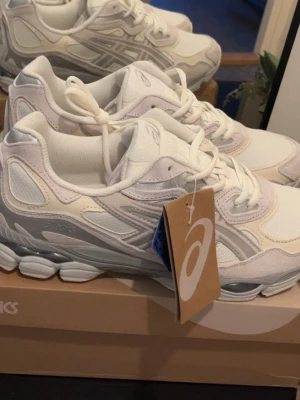 Asics GEL NYC - Snygga vita sneakers från ASICS med grå och ljusbeige detaljer. Skorna har en chunky sula och klassisk mesh- och syntetöverdel för skön komfort och sportig look. Perfekta för dig som gillar stilrena och trendiga sneakers.