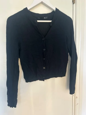 Svart kofta från 157 med knappar - Klassisk svart kofta från 157 med rund halsringning och knäppning framtill. Långärmad modell med ribbade muddar vid ärmslut och nederkant. Perfekt att slänga över en t-shirt eller linne för en enkel och clean look.