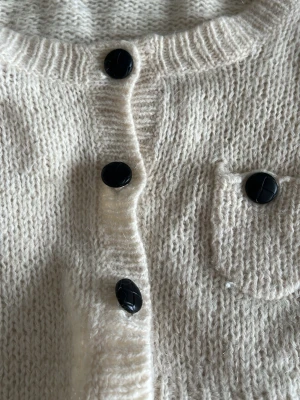 Krämvit kofta från Filippa K - Mysig krämvit kofta från Filippa K med stora svarta knappar och en liten bröstficka. Koftan är stickad i en mjuk blandning av mohair, polyamid, lammull och angora. Perfekt för lager på lager och ger en clean, stilren look.