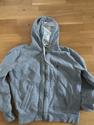 Grå hoodie från Polo Ralph Lauren - Klassisk grå hoodie med dragkedja från Polo Ralph Lauren. Tröjan har huva med vit dragsko, två fickor framtill och den ikoniska lilla mörkblå loggan broderad på bröstet. Tillverkad i mjuk bomullsblandning som är skön att ha på sig.