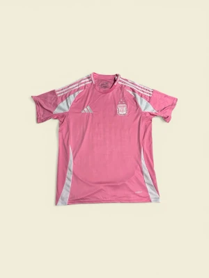 Rosa Argentina Adidas Jersey☀️ - Snygg rosa träningströja från Adidas med Argentinas fotbollsförbunds logga på bröstet. Tröjan har vita detaljer och klassiska Adidas-ränder på axlarna. Tillverkad i lätt och ventilerande funktionsmaterial, perfekt för träning eller sport.